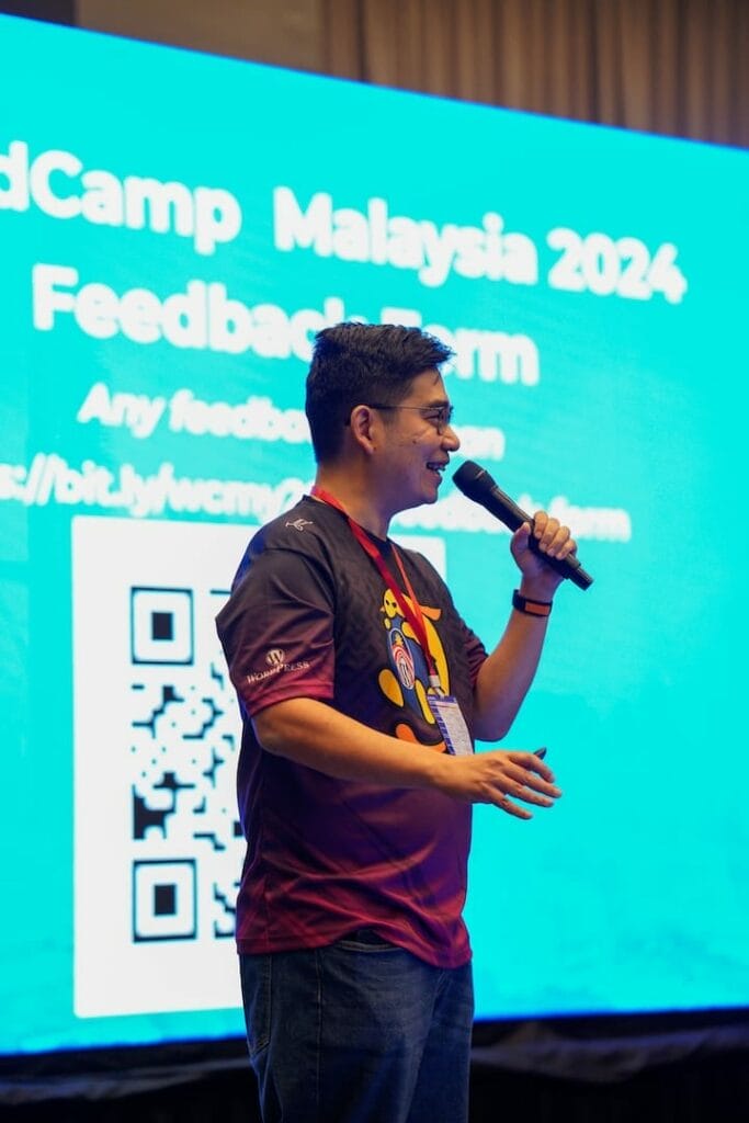 A person speaks into a microphone in front of a blue screen displaying "WordCamp Malaysia 2024 Feedback Form" with a QR code and a feedback link, seamlessly addressing questions about Leokoo.